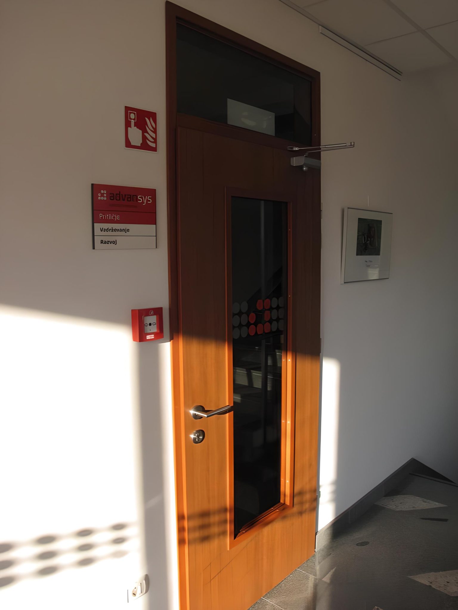 Lesena notranja vrata s steklenim panelom v poslovnem objektu Advansys s tablo oddelkov ob njih.